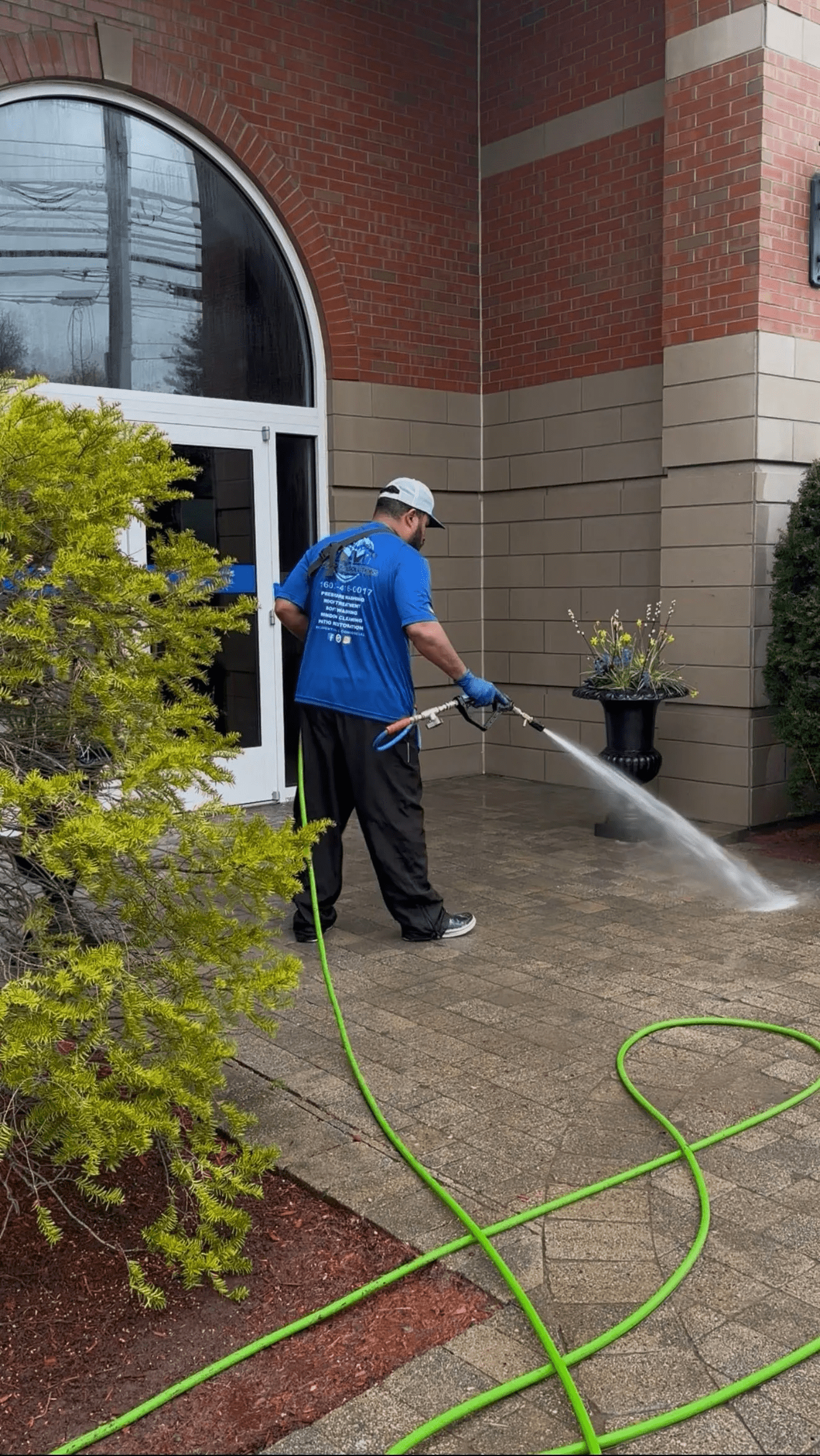 A technician pressure washing a brick paver walkway and plaza area in front of a commercial building entrance.