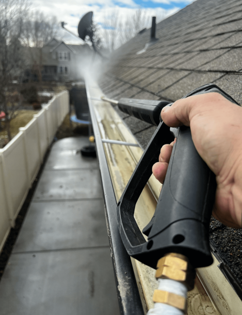 Close-up first-person view of a technician using a pressure washer spray wand to clean out a residential gutter line.